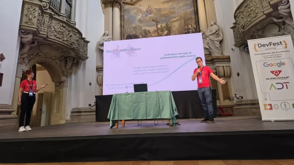 Isabella Flecchia and Gaetano Castaldo on stage at DevFest Modena 2026, inside the church of Fondazione San Carlo, in front of the slide with the three questions on professional identity